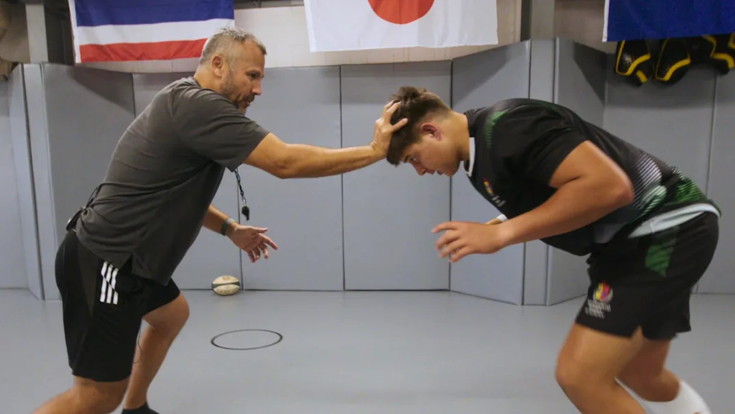 Coach working through contact body-shape technique with a young rugby player on a contact-pad surface, framed by team flags overhead — example of the kind of training footage a program uploads into Elite Rugby App.